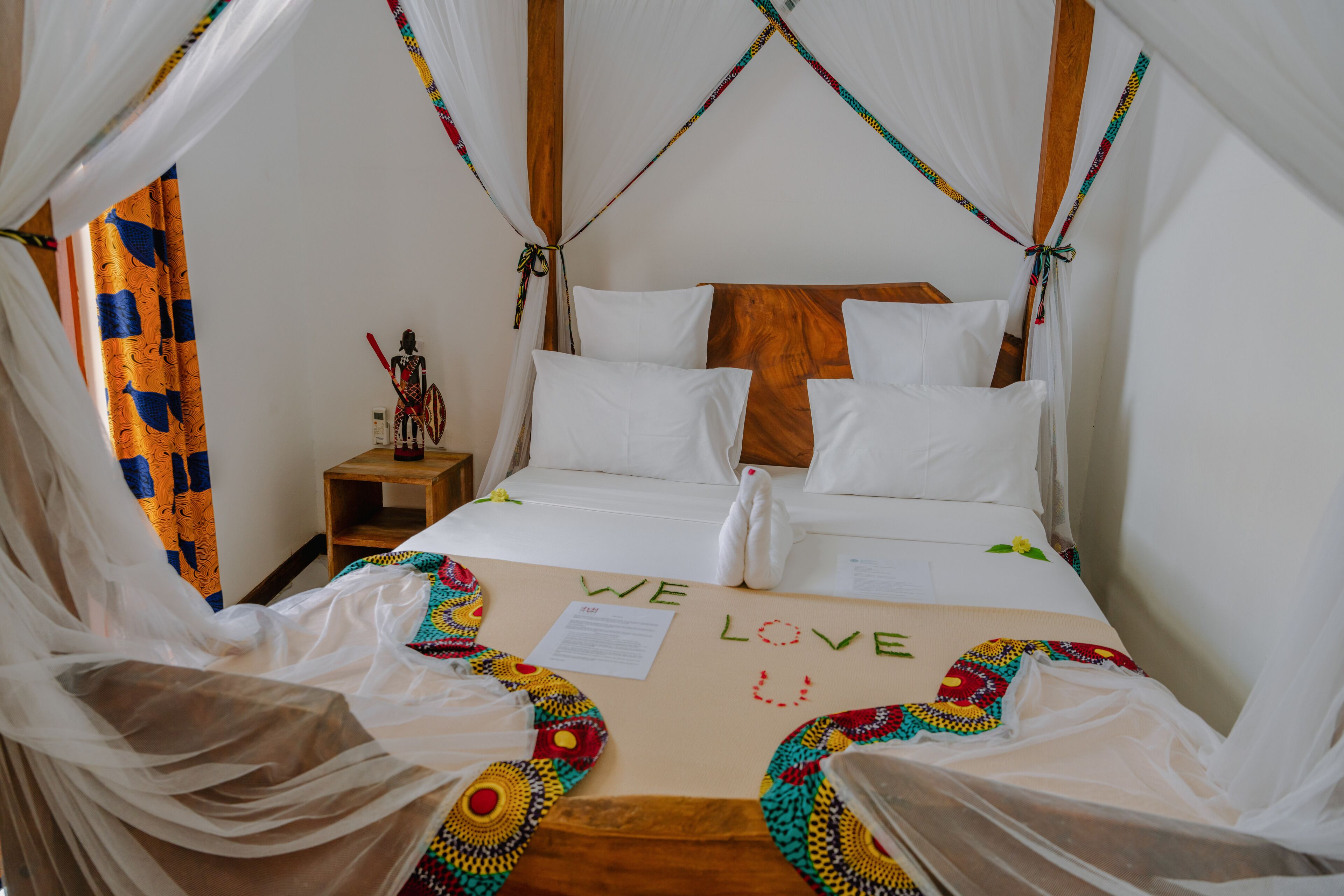 A bed with white linens, a wooden headboard, and a canopy. There is a note on the bed that says "WE LOVE YOU".