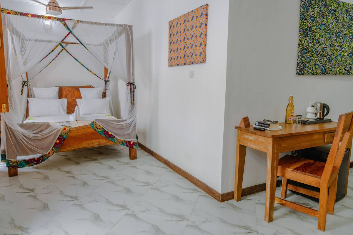 Une chambre avec un lit sous un dais, un bureau en bois avec une chaise, et un mur orné de deux œuvres d’art.