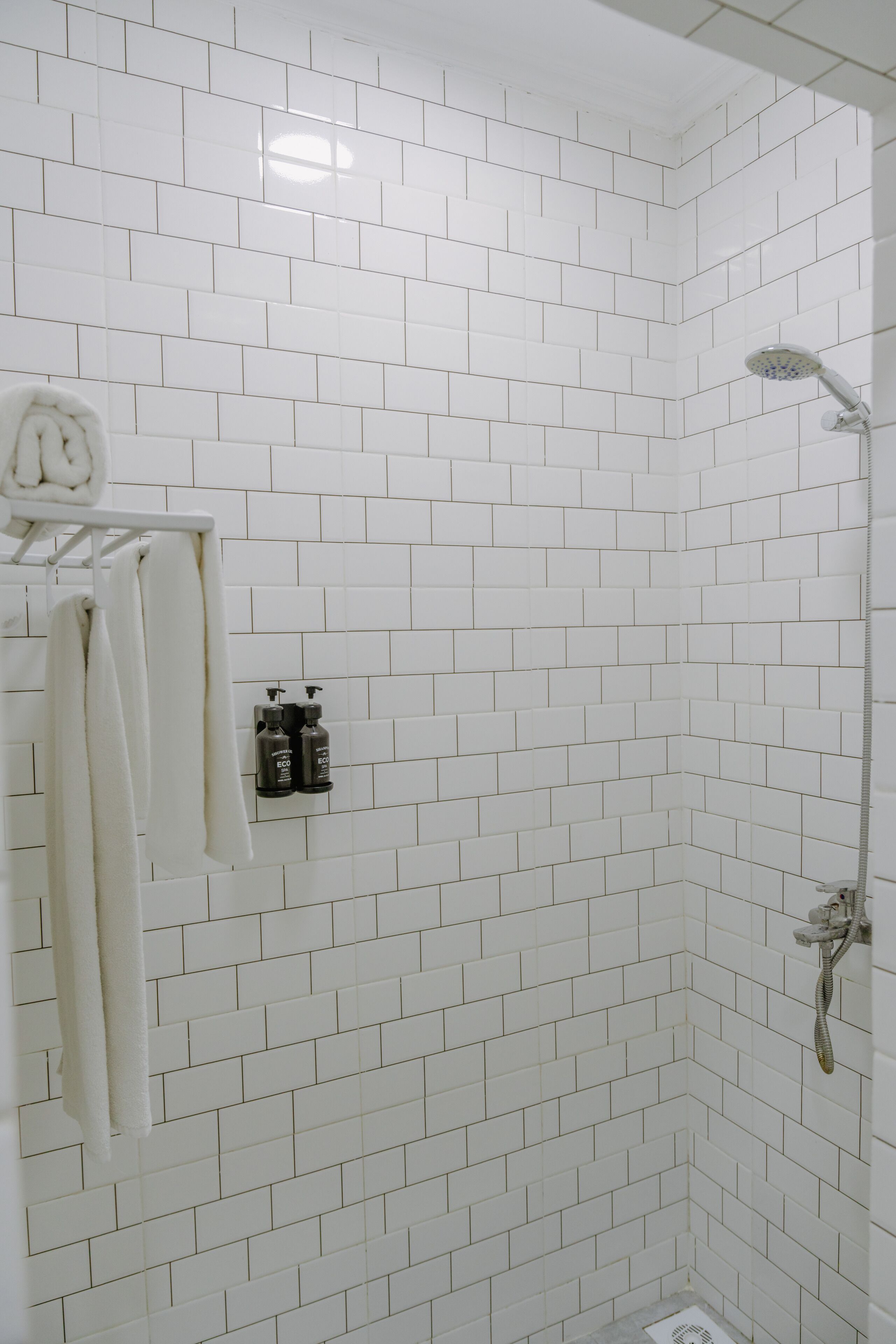 Een badkamer met witte betegelde wanden en een douche. Twee flessen shampoo en conditioner hangen aan de muur.