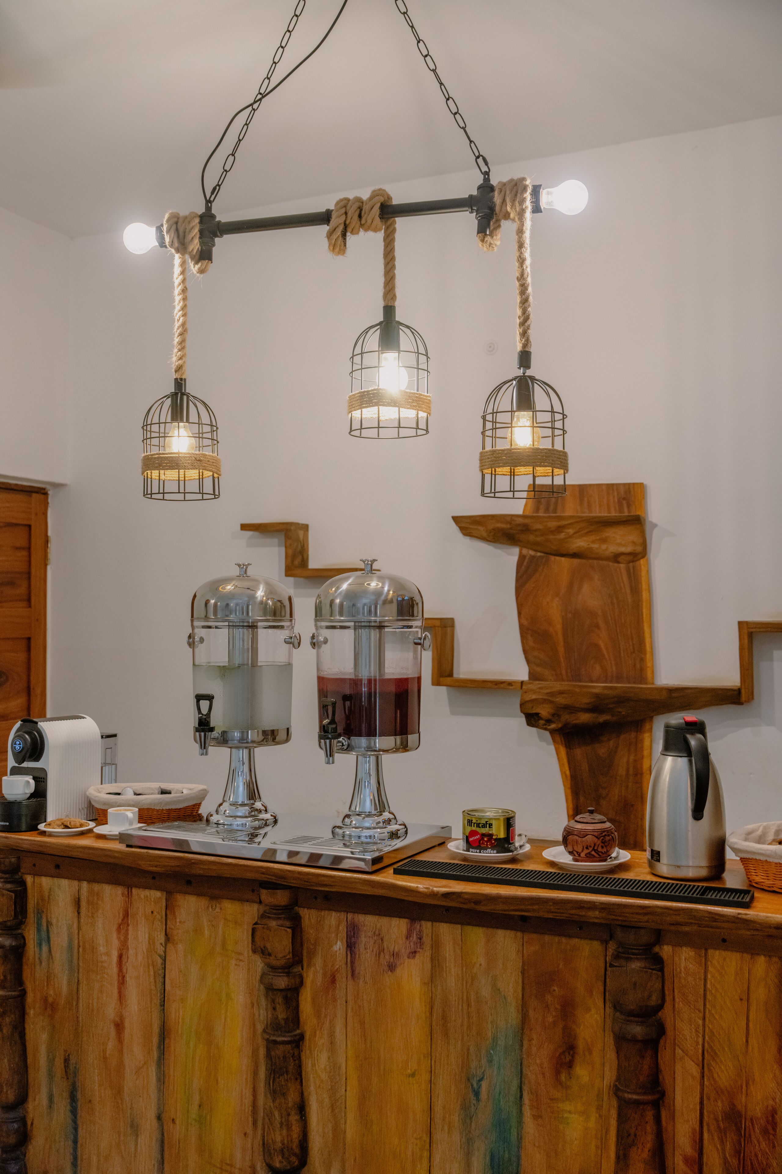 Uma área de bar com balcão de madeira, dois grandes dispensadores de água, uma máquina de café e uma chaleira.