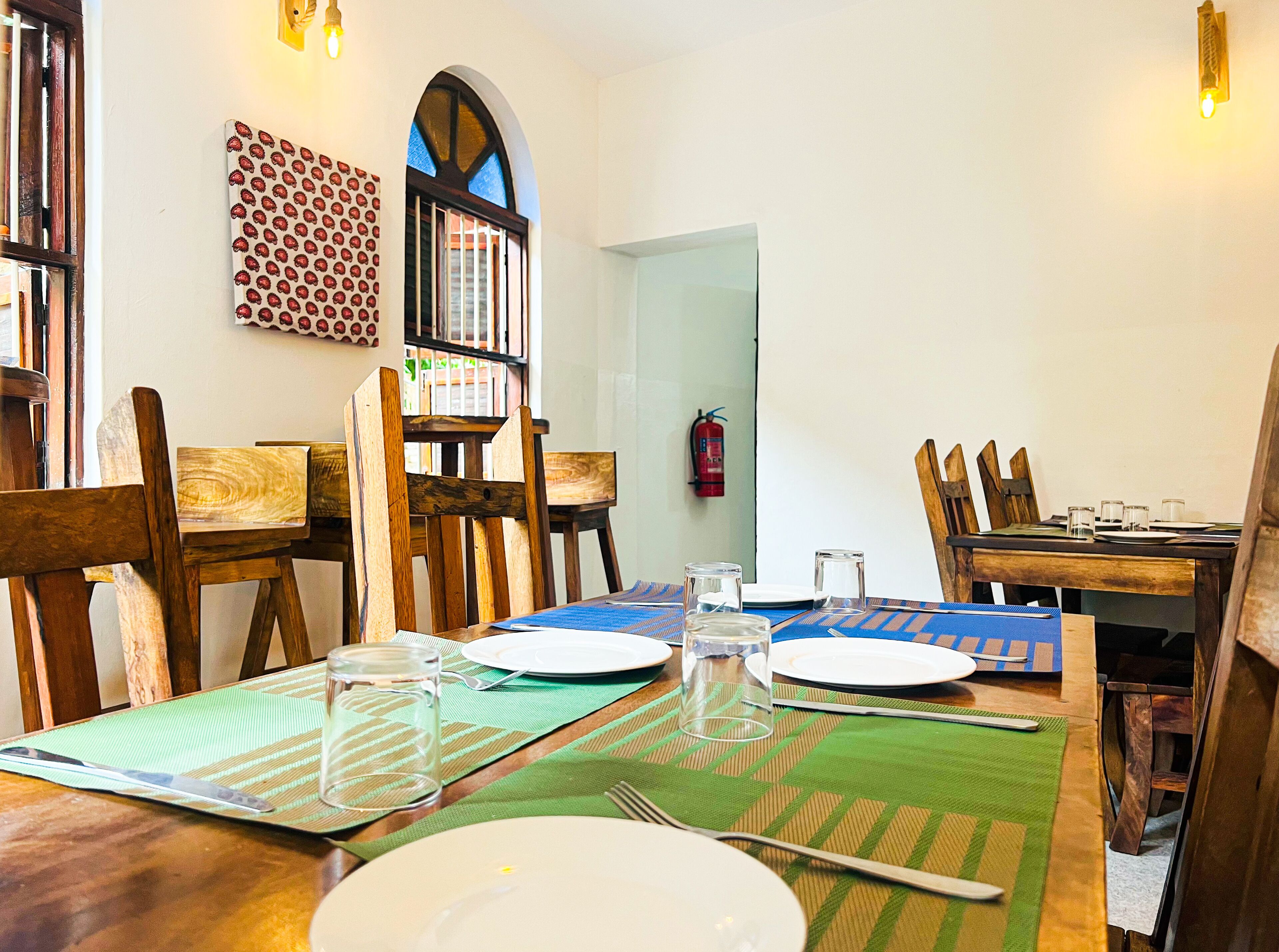 A dining area with wooden chairs and tables, a fire extinguisher on the wall, and a window with a decorative pattern.