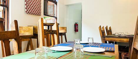 A dining area with wooden chairs and tables, a fire extinguisher on the wall, and a window with a decorative pattern.