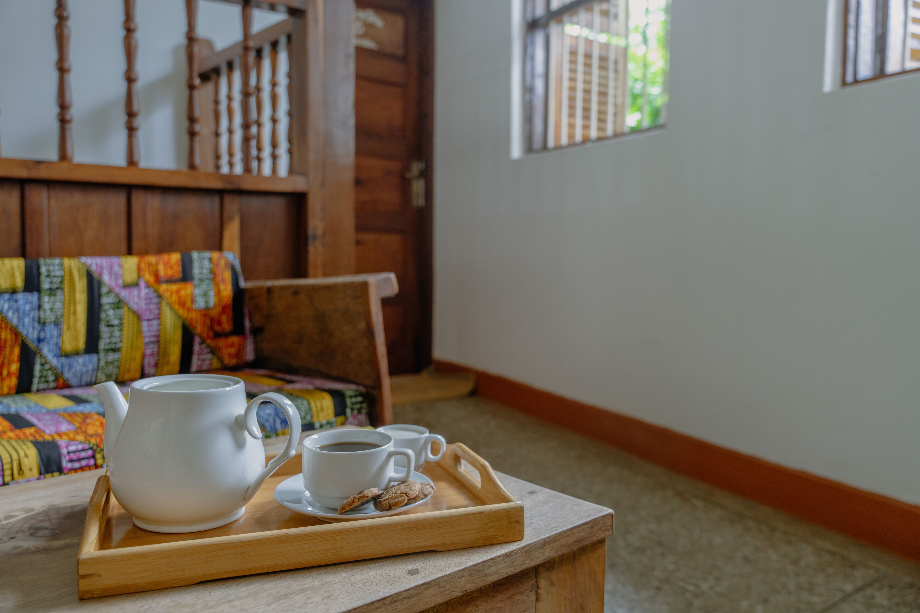 A wooden tray with a teapot, cups, and cookies on a table in a room with wooden furniture and a window.