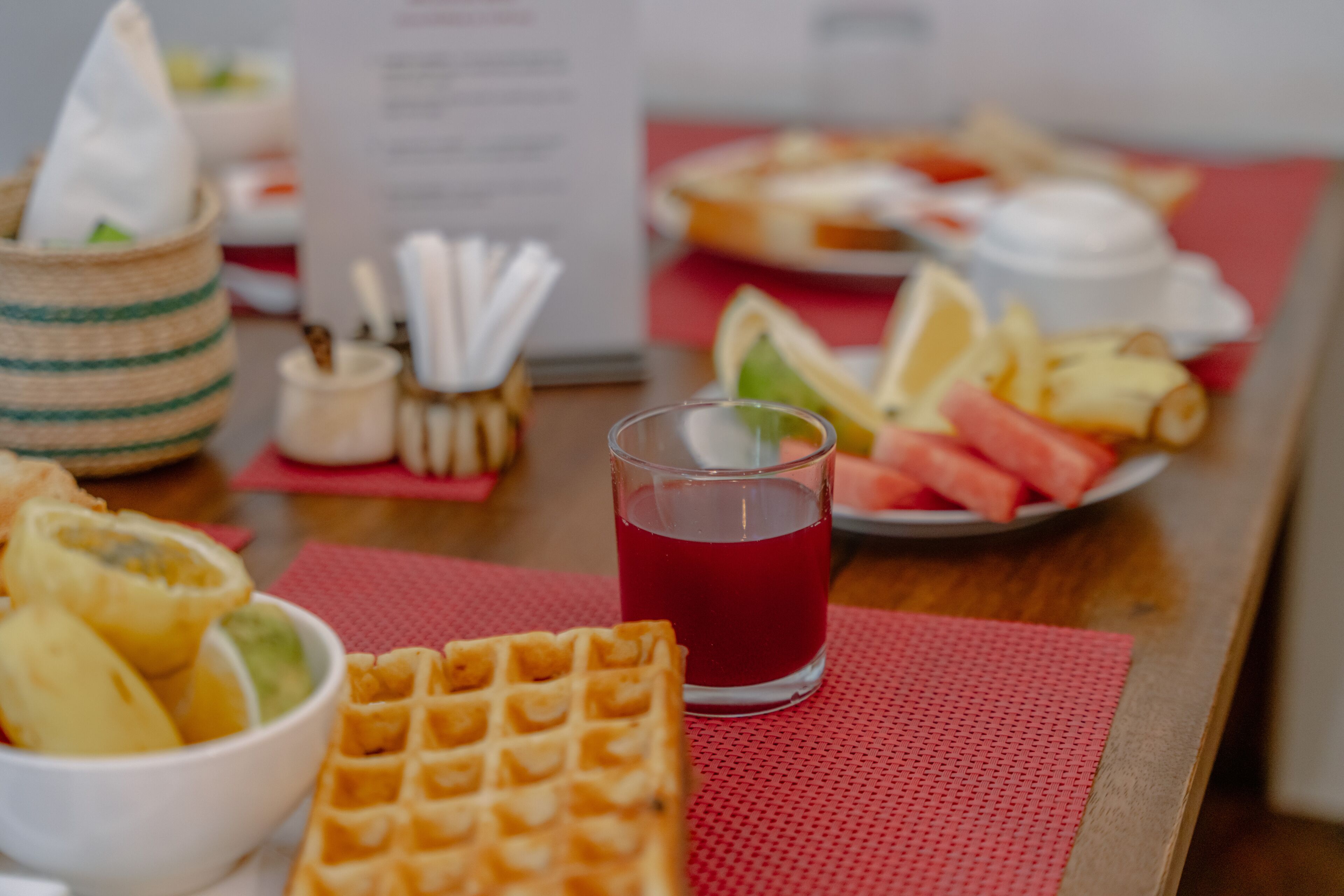 A breakfast setting with a waffle, fruit bowl, and a glass of red beverage on a red placemat.