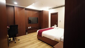 A modern hotel room with a wooden floor, a bed with a red and white bedspread, a flat-screen TV mounted on the wall, a desk with a chair, and a trash bin.