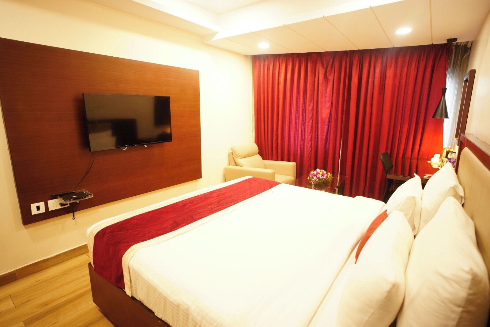 A hotel room with a large bed, a TV mounted on the wall, a red curtain, and a small table with a vase of flowers.