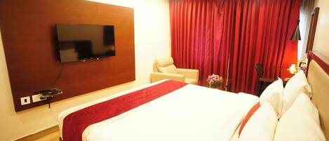 A hotel room with a large bed, a TV mounted on the wall, a red curtain, and a small table with a vase of flowers.