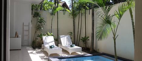 A swimming pool area with two white lounge chairs, a small white table, and a ladder against a wall.