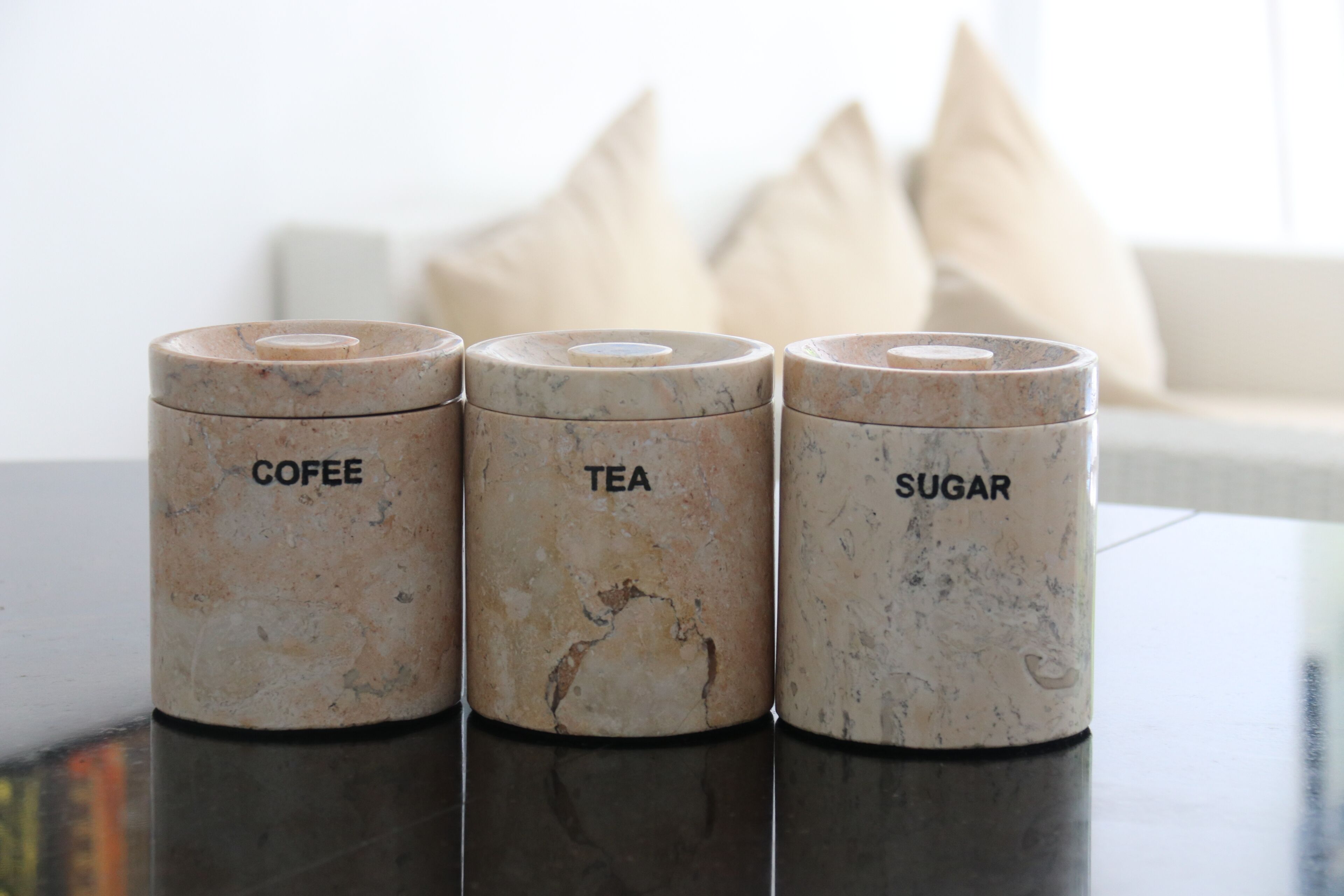 Tres recipientes de cerámica con las etiquetas CAFÉ, TÉ y AZÚCAR sobre una superficie reflectante.