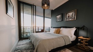 Un dormitorio con un ventanal grande, una cama con colcha estampada, una mesita de noche con lámpara y una pila de libros.