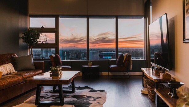 Una sala de estar moderna con un sofá de cuero, una mesa de centro de madera y un ventanal con vistas a la ciudad al atardecer.