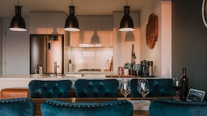 Una cocina moderna con iluminación colgante, una isla central y una zona de comedor con sillas tapizadas en color verde azulado.