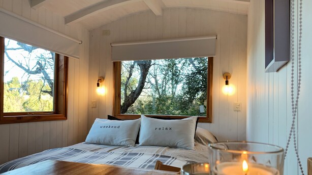 Een gezellige slaapkamer met een groot bed, twee kussens met tekst, een houten tafeltje met een kaars en uitzicht op bomen door een raam.