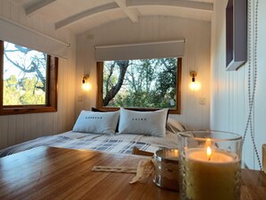Un dormitorio acogedor con una cama grande, dos almohadas con texto, una mesa de madera con una vela y vistas a árboles a través de una ventana.