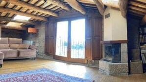 Una habitación rústica con pared de piedra, vigas de madera, un sofá, chimenea y un ventanal grande.