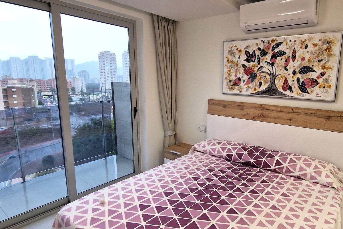 A hotel room with a large window, a bed with a geometric patterned bedspread, a wooden headboard, and a wall-mounted air conditioner.