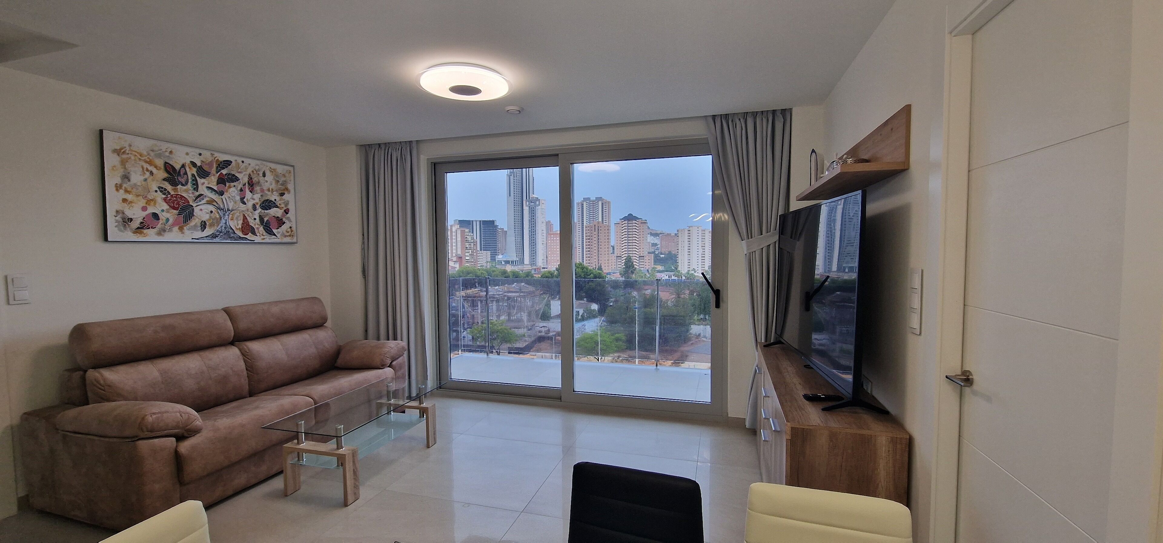 A modern living room with a brown sofa, a glass coffee table, a flat-screen TV on a wooden stand, and a large window with curtains.
