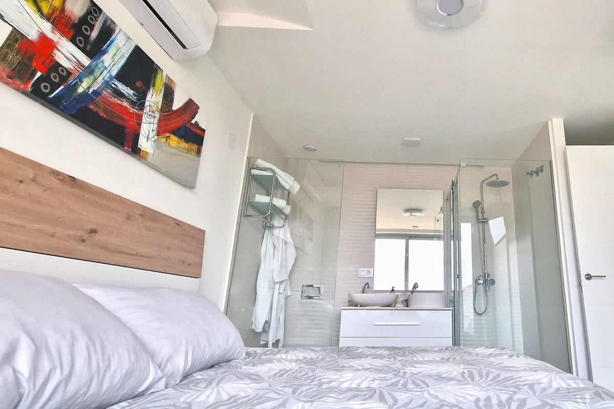 A modern bedroom with a bed, a bedside table, a mirror, a shower, and a TV.