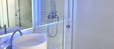 Un baño moderno con ducha de vidrio, un lavabo blanco y encimera de mármol, y un espejo grande.