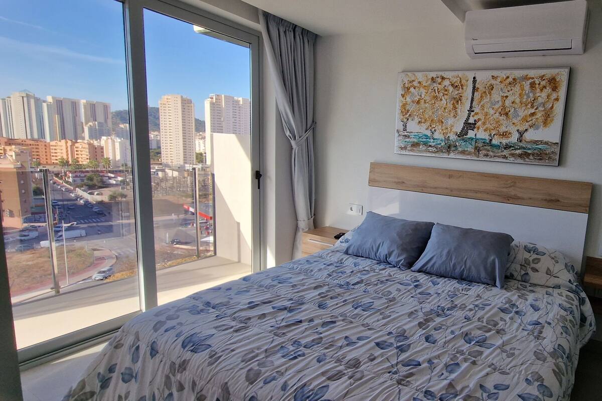 A bedroom with a large window, a bed with floral bedding, a headboard, a painting of trees, and a glass shower area.