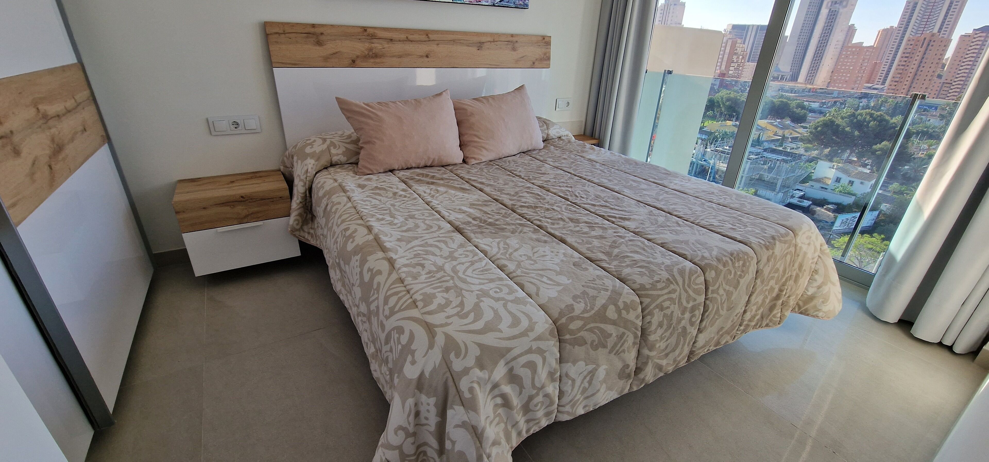 Une chambre moderne avec un grand lit, une tête de lit en bois et une vue sur le paysage urbain grâce à de grandes fenêtres du sol au plafond.