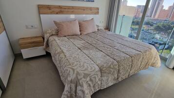 Une chambre moderne avec un grand lit, une tête de lit en bois et une vue sur le paysage urbain grâce à de grandes fenêtres du sol au plafond.