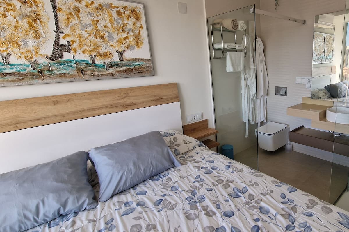 A bedroom with a bed, a wooden headboard, a painting, a shower, and a glass-enclosed bathroom with a mirror.