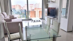 A modern living room with a glass dining table, white sofas, and a large window with a city view.
