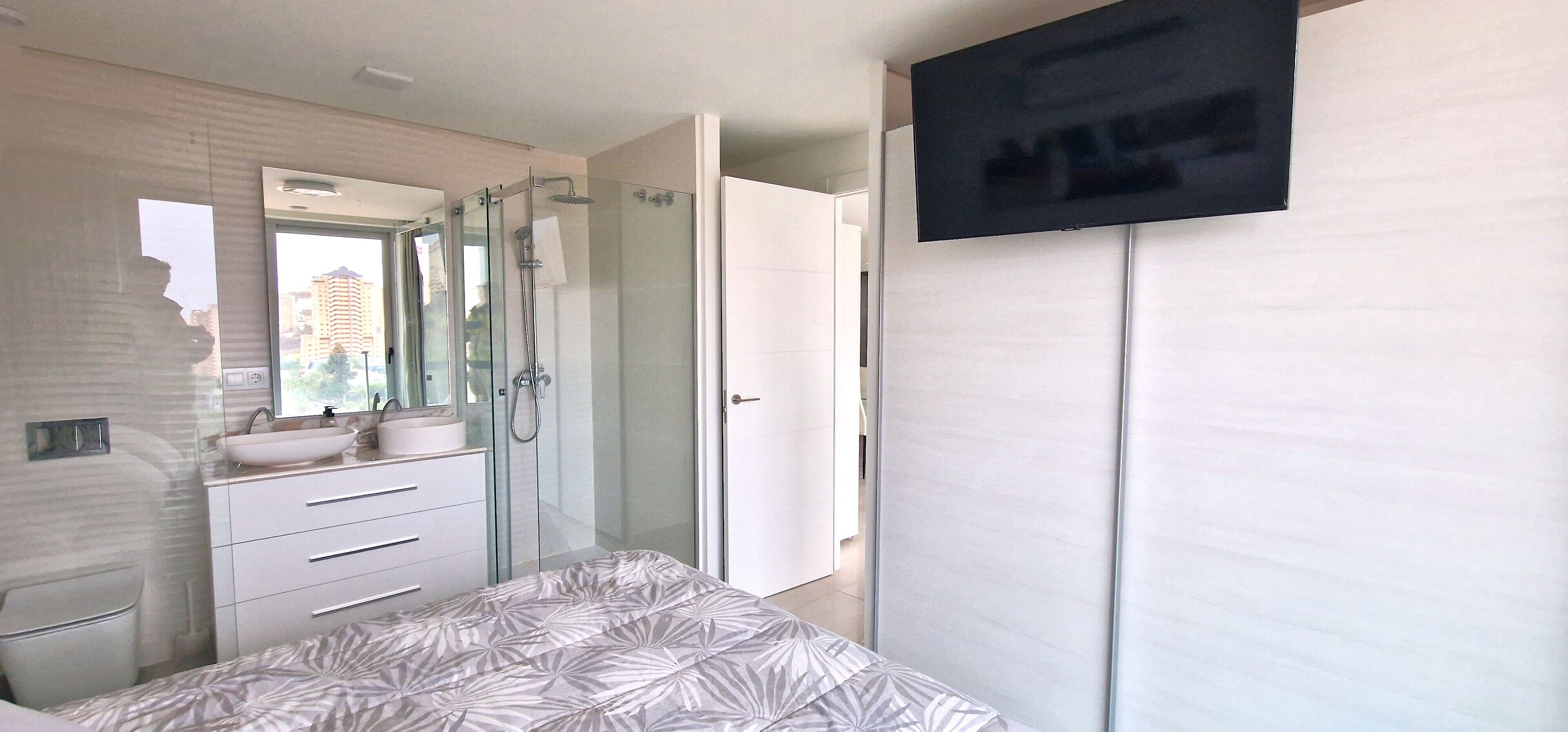 A modern bedroom with a large bed, a glass-enclosed shower, a white vanity with a sink, and a flat-screen TV mounted on the wall.