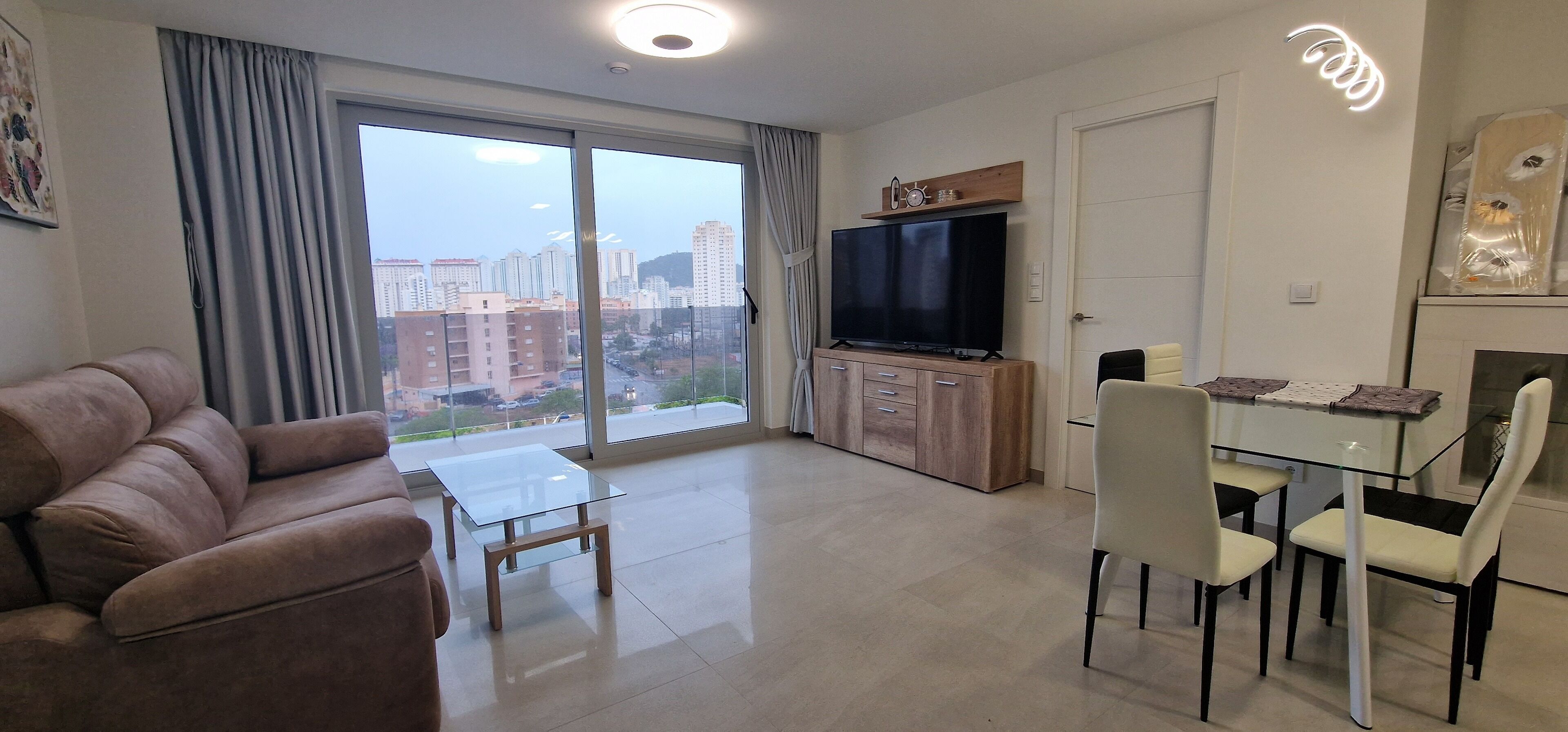 Una sala moderna con sofá, una mesa de centro de vidrio, un televisor sobre un mueble de madera y una zona de comedor con sillas y una mesa.