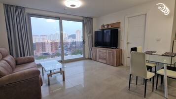 Una sala moderna con sofá, una mesa de centro de vidrio, un televisor sobre un mueble de madera y una zona de comedor con sillas y una mesa.