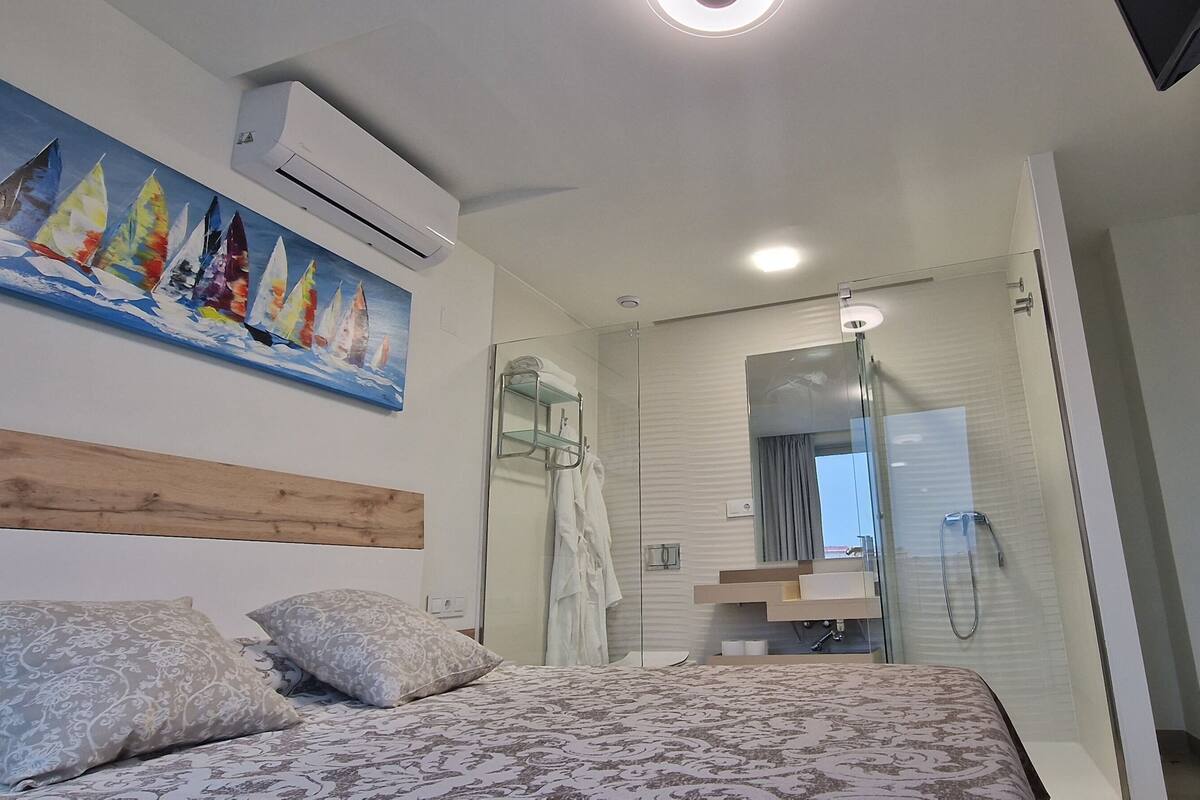 A modern bedroom with a bed, patterned pillows, a painting of sailboats, a glass shower enclosure, and a wooden headboard.