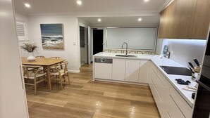 A modern kitchen with white cabinetry, a wooden dining table, and a view of the ocean through a large window. - Cozy House in Forresters Beach - modern interiors  (Forresters Beach)