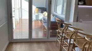 A modern kitchen with wooden flooring, a dining area with a table and chairs, and a view through large sliding glass doors to an outdoor deck. - Cozy House in Forresters Beach - modern interiors  (Forresters Beach)