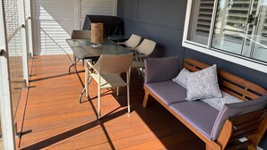 A covered patio with a glass table, wicker chairs, a wooden bench, and a window with blinds. - Cozy House in Forresters Beach - modern interiors  (Forresters Beach)