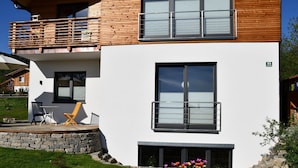 A modern two-story building with a balcony, a stone wall, and a small outdoor seating area with chairs. - New 2 room apartment in Murnauer Moos (Schwaigen)