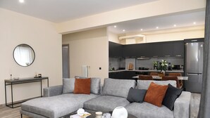 Uma sala de estar moderna com um sofá modular cinza, uma mesa de centro redonda e uma pequena mesa lateral. Uma cozinha é visível ao fundo, com armários escuros e eletrodomésticos modernos.