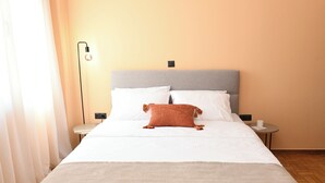 Cama bem arrumada com lençóis brancos e um único travesseiro marrom, mesa de cabeceira com abajur e pequena mesa com xícara e cardápio.