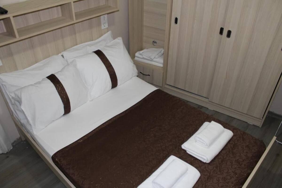 A neatly made bed with white linens and a brown blanket, a small bedside table with a folded towel, and a wooden wardrobe with two doors.