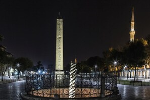 Un monument orné d’inscriptions, entouré d’une grille décorative et donnant sur une place dallée, illuminée par des lampadaires.