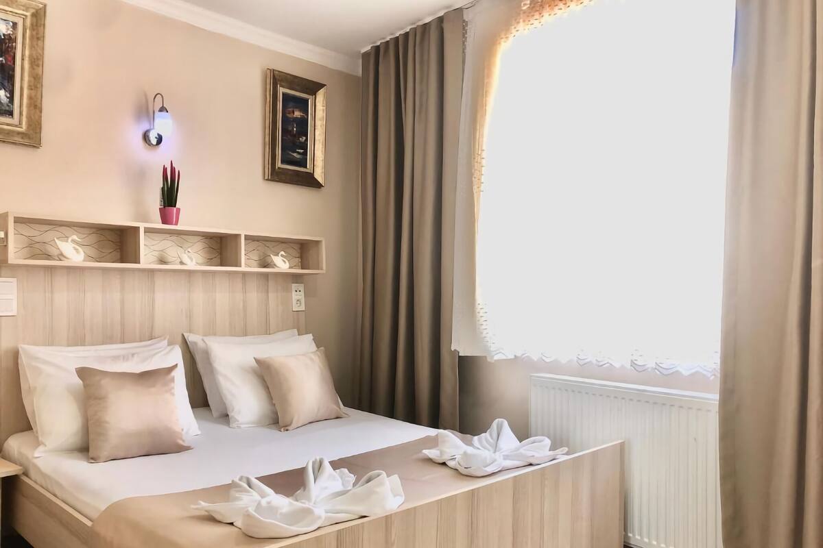 A hotel room with a bed, a bedside table, a nightstand, a wall-mounted shelf with decorative items, and a window with curtains.