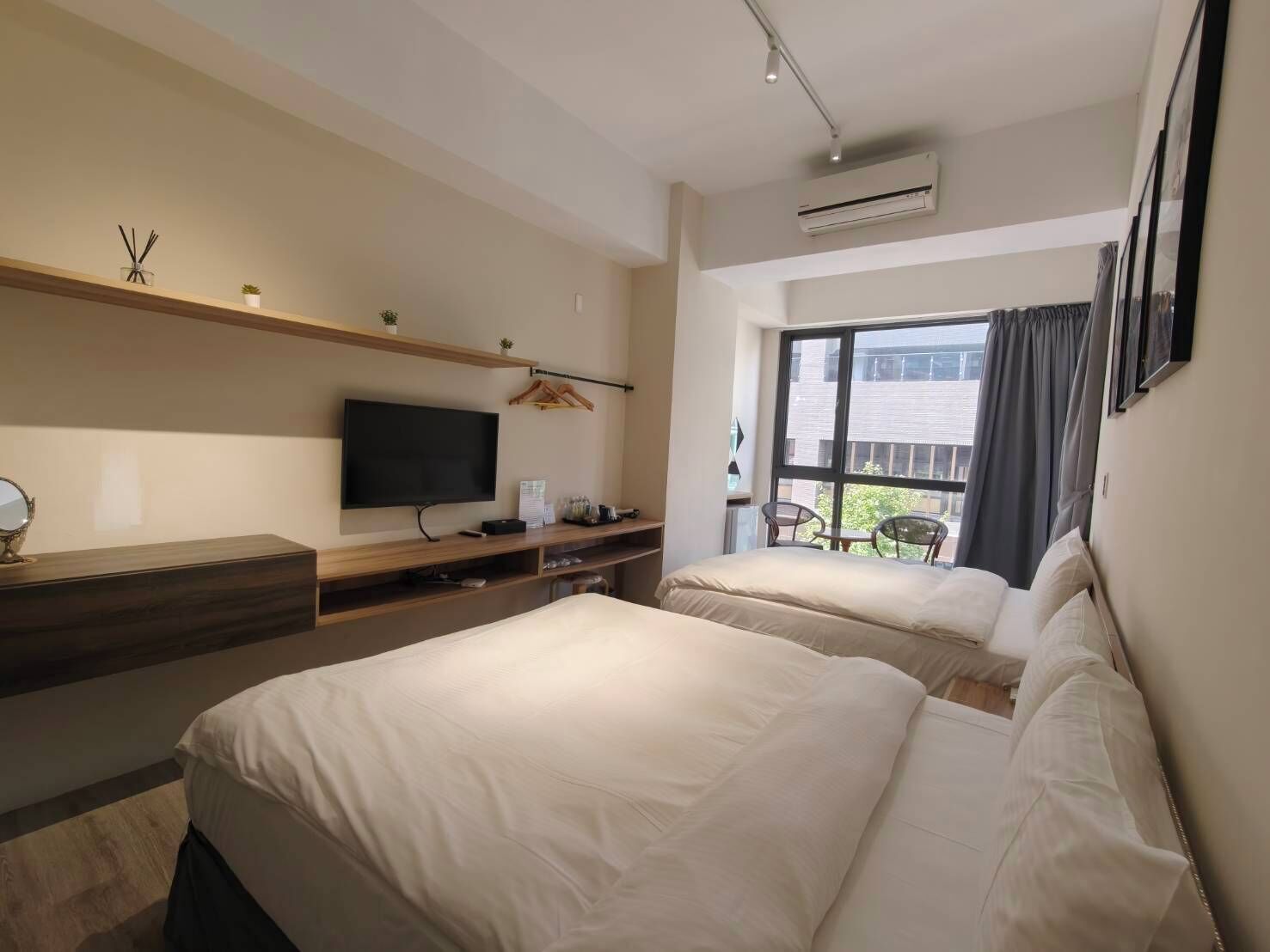 A modern hotel room with two beds, a TV, a desk, and a balcony with a view of the city.