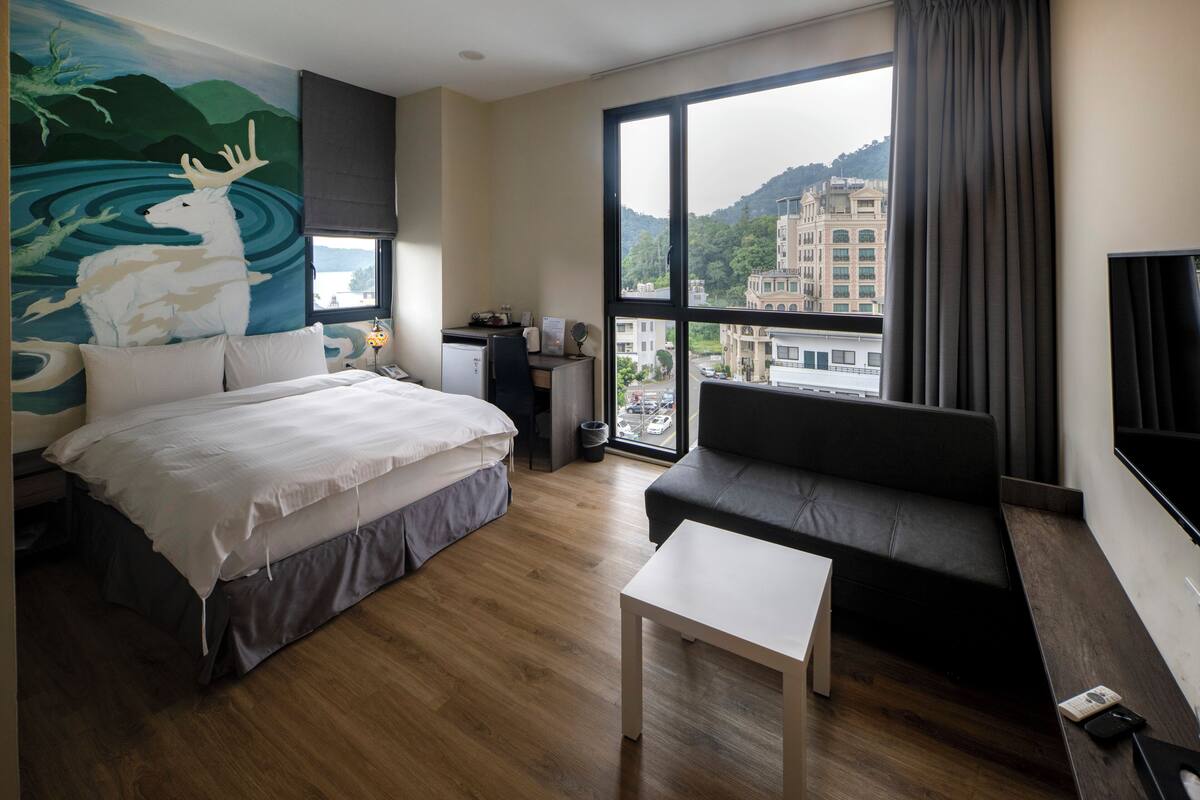Ett hotellrum med en säng, en soffa, ett litet bord, ett skrivbord och ett stort fönster med utsikt över staden.