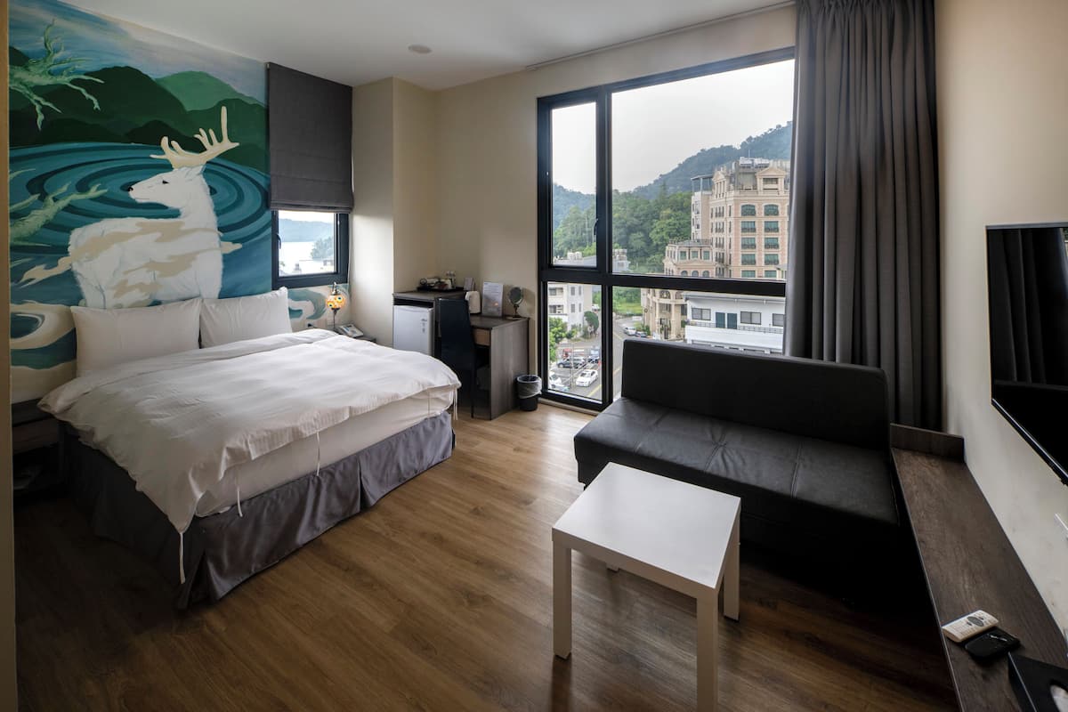 A hotel room with a bed, a sofa, a small table, a desk, and a large window offering a city view.