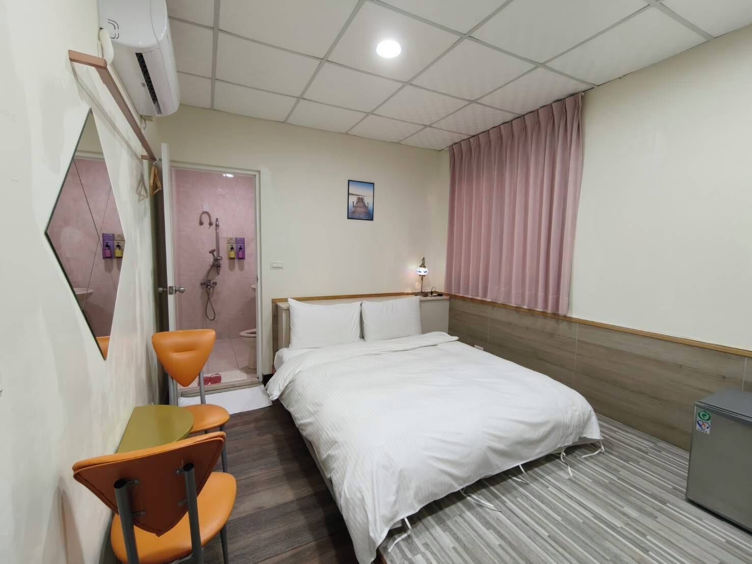 A hotel room with a bed, a desk with a chair, a shower area, and a small refrigerator.