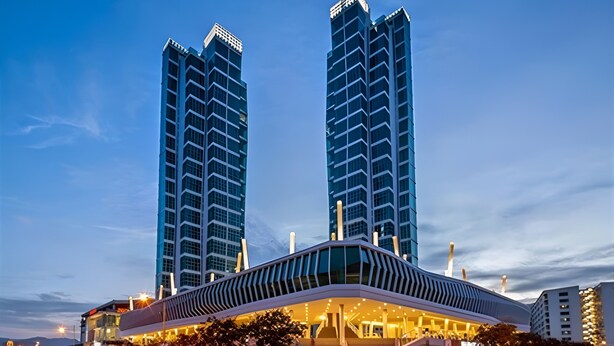 A modern hotel with a curved facade, two tall adjacent buildings, and a parking area with cars.
