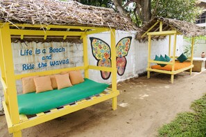 Un área de estar al aire libre con temática playera, que incluye un mural, techo de paja y suelo de arena.