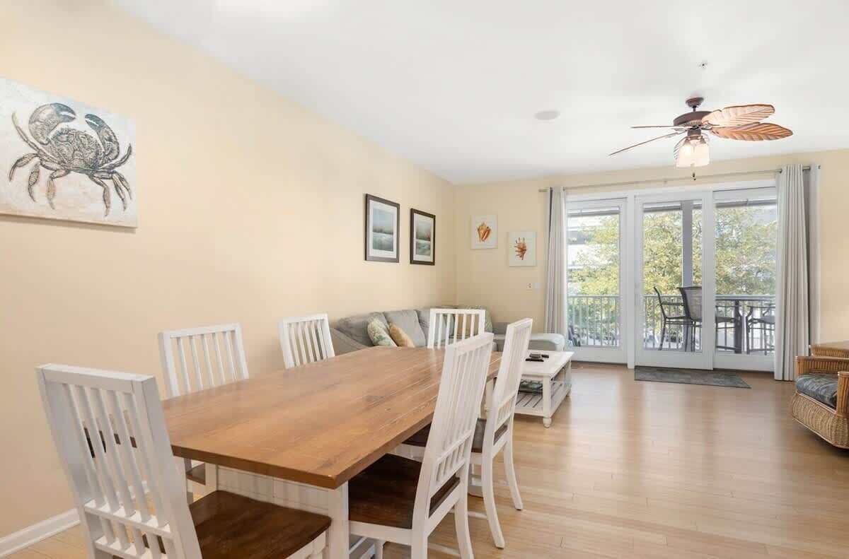 A dining area with a wooden table and white chairs, a ceiling fan, and a view of the outdoors through large sliding glass doors.