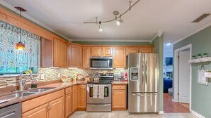 Une cuisine moderne avec des meubles en bois, des appareils en acier inoxydable, un four à micro-ondes, un four et un réfrigérateur. Il y a une fenêtre avec un rideau décoratif et un luminaire suspendu.
