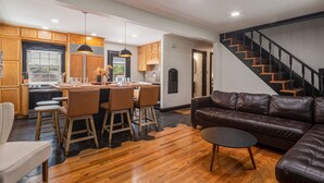 A modern kitchen with wooden cabinets, a dining area with bar stools, a leather sofa, and a staircase. - ꕥLuxꕥHeated* Pool/Hot Tub/Gamerooms/Firepit/Outdoor Shower/Bunks (New Albany)